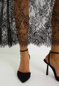 Superposition de dentelle noire avec des motifs floraux, associée à des talons noirs pointus et des brides à la cheville, sur un fond neutre.