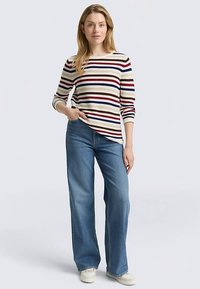 Gestreiftes Langarmshirt in Creme mit roten, blauen und braunen Streifen; dazu hellblaue Jeans mit weitem Bein und weiße Sneaker.