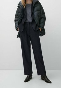 Kvinde iført en sort dunjakke over en mørk blazer og en grå sweater, kombineret med sort brede bukser og sorte ankelstøvler.