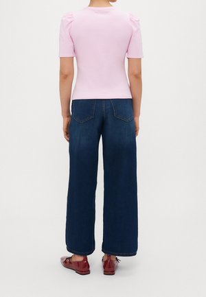 Persona con camiseta rosa clara de manga corta, vaqueros oscuros de pierna ancha y zapatos de charol rojos, de espaldas.