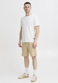 White cotton t-shirt, beige shorts, beige sneakers with stripes, white socks, standing against a plain background. Minimalist design, casual look.