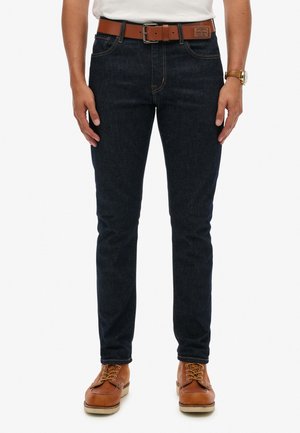 Marineblå denimjeans med smal pasform, synlige sømme og et læderbælte. Båret med brune sko og udstråler et afslappet look.