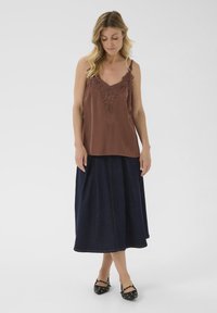 Camisole marron avec une bordure en dentelle, associé à une jupe en denim fluide de couleur foncée. Le mannequin porte des ballerines noires avec des accents argentés. Fond minimaliste.