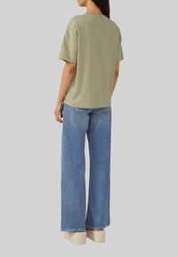 T-shirt oversize vert olive à manches courtes, associé à un jean large bleu clair, avec une coupe décontractée et un branding minimal.