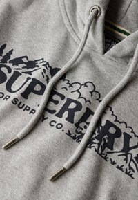 Grijze hoodie met een groot zwart logo en berggraphics, voorzien van de tekst "Superdry". Heeft trekkoorden met metalen uiteinden en een gestructureerd oppervlak.