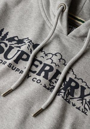 Felpa grigia con cappuccio caratterizzata da un grande logo nero con grafiche di montagne e la scritta "Superdry". Dotata di lacci con punte in metallo e una superficie testurizzata.