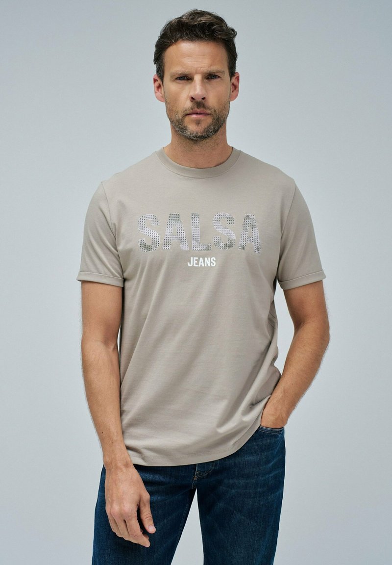 Světle béžové tričko s krátkým rukávem, na kterém je nápis "SALSA JEANS" ve rhinestonech napříč hrudníkem, v kombinaci s tmavými denimovými džíny.