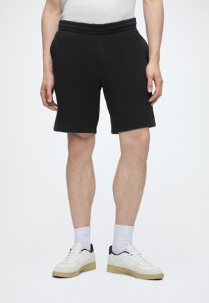 Homme portant un short noir à taille élastique, des chaussettes blanches et des baskets blanches avec des semelles beige sur un fond clair uni.