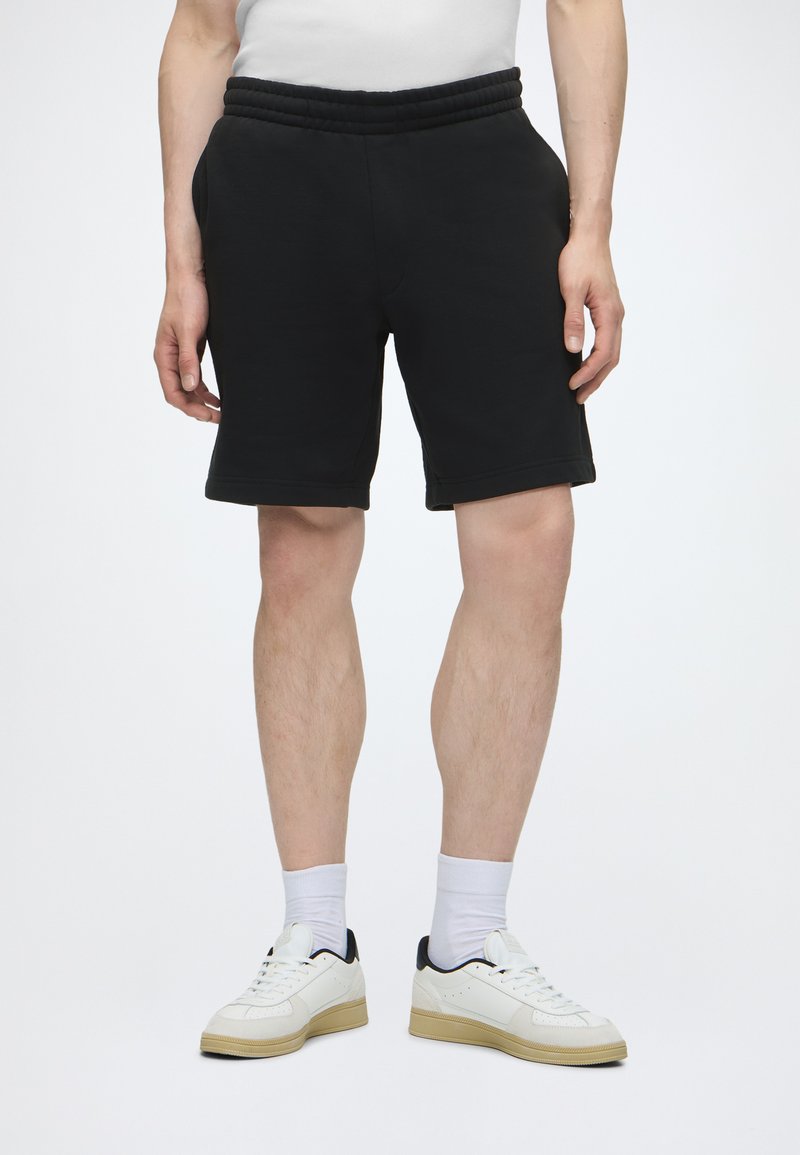 Homme portant un short noir à taille élastique, des chaussettes blanches et des baskets blanches avec des semelles beige sur un fond clair uni.