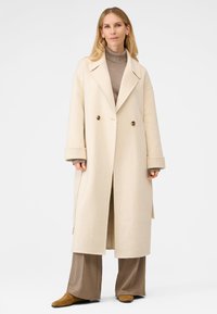 Cremefarvet dobbeltradet frakke med store reverser, lavet af blødt stof. Båret over en brun turtleneck, sammen med brede brune bukser og brune sko.