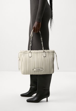 Persona in abito grigio a coste e stivali neri con tacco che tiene una grande borsa beige pieghettata con manici a catena argentata.