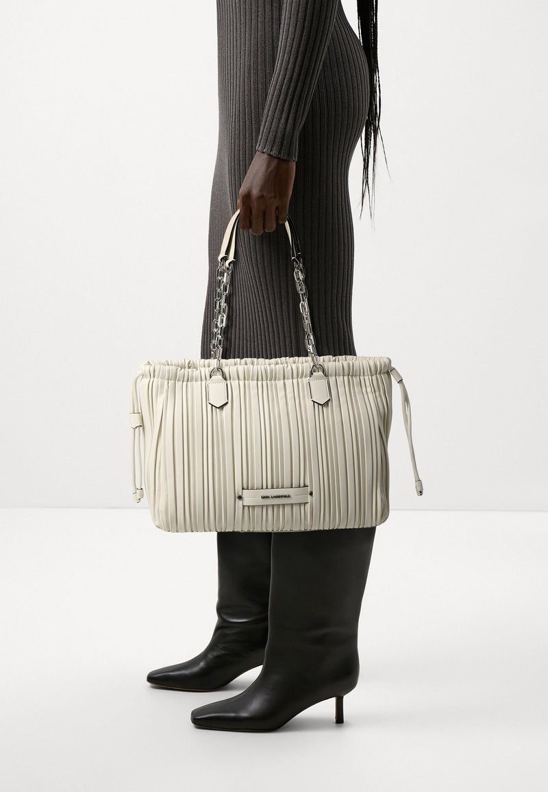 Persoon in een geribbelde grijze jurk en zwarte laarzen met hak, die een grote beige geplooide handtas met zilveren kettinghandvatten vasthoudt.