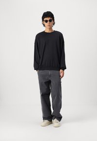 Black oversized sweatshirt with dropped shoulders, paired with loose, gray pants. Model wears round, dark sunglasses and beige shoes.