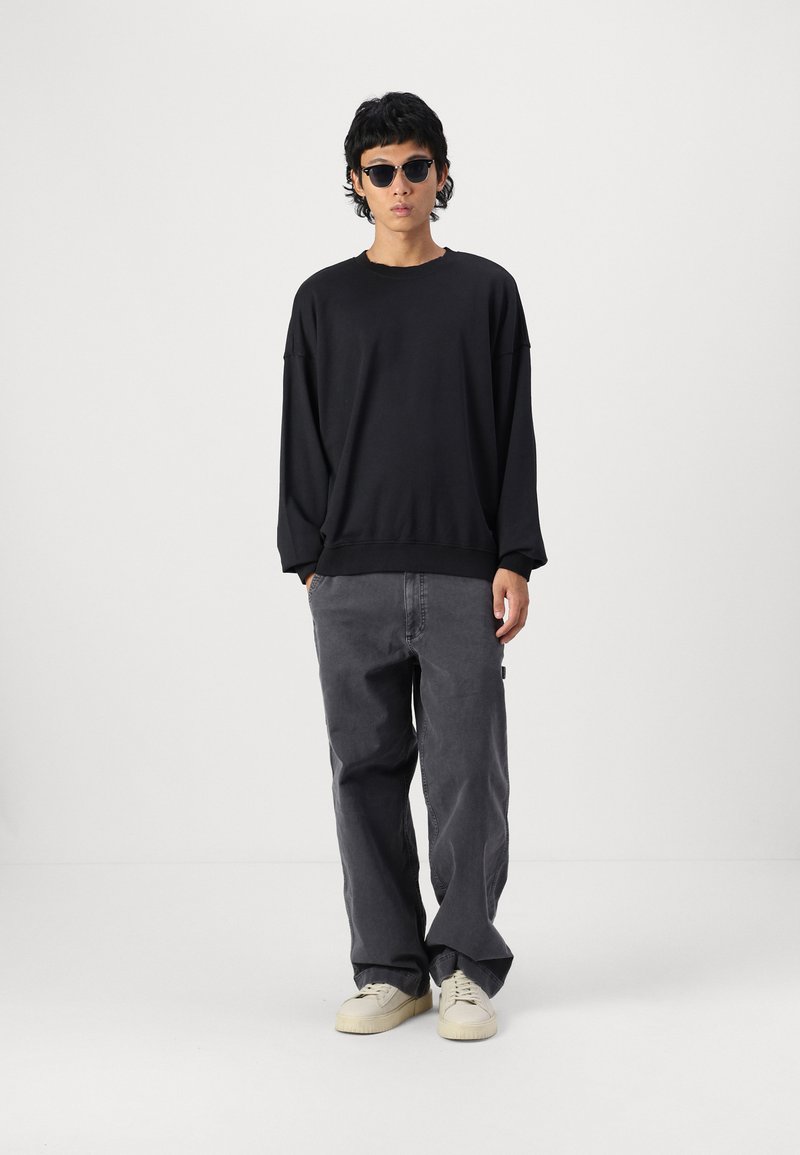 Black oversized sweatshirt with dropped shoulders, paired with loose, gray pants. Model wears round, dark sunglasses and beige shoes.