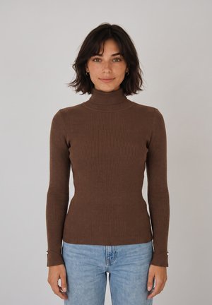 Donna con capelli corti e scuri che indossa un maglione marrone a coste con collo alto e jeans azzurri, in piedi davanti a uno sfondo chiaro e uniforme.