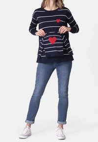 Pruhovaný tmavě modrý svetr s červeným detailem ve tvaru srdce, doplněný džínami a bílými teniskami. Svetr obsahuje uvnitř odpovídající mini svetr.