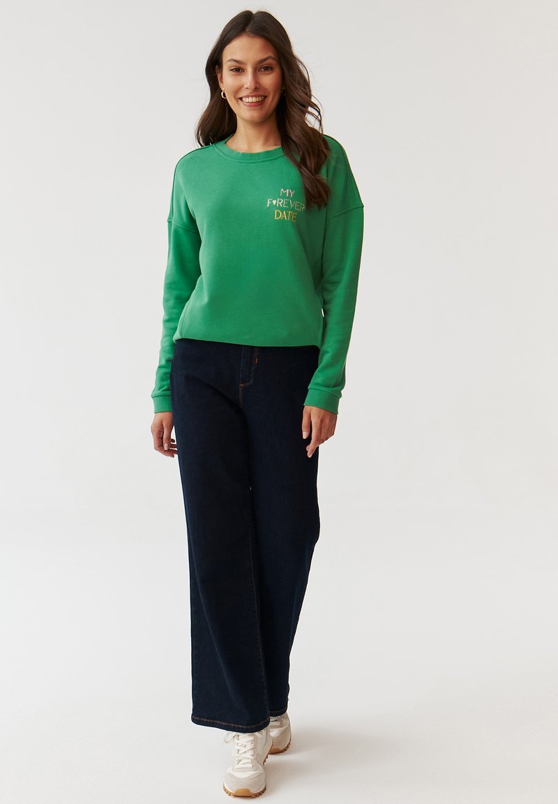 Zielona bluza z nadrukiem "MOJA NA ZAWSZE RANDKA" w kolorze żółtym, zestawiona z ciemnoniebieskimi szerokimi spodniami i białymi trampkami. Gładka faktura.