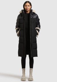 Hosszú fekete kabát steppelt panelekkel, bézs színű szőrmével bélelt kapucnival, elöl cipzáras, oldalán zsebekkel. Egy fekete testhezálló ruházat és szürke csizmák fölött viselve.