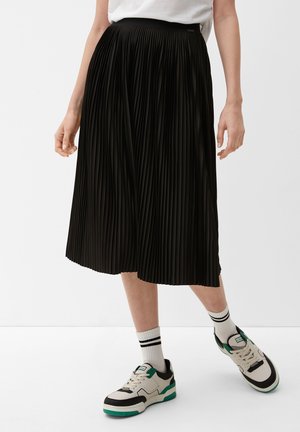 Zwarte geplooide midi-rok met een hoge taille en een gestructureerde stof, gecombineerd met witte en groene sneakers en enkelsokken met twee zwarte strepen.