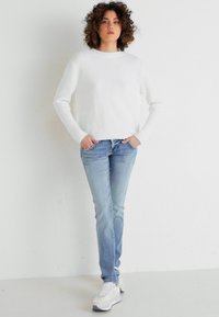 Vit rippad tröja, ljusblå jeans och vita sneakers. Modellen står vänd mot kameran med lockigt hår, händerna i fickorna mot en neutral bakgrund.