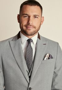 Traje gris con solapas en punta, acentos marrones en contraste, camisa blanca, corbata de lunares y pañuelo de bolsillo con patrón en el bolsillo del pecho.