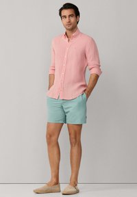 Hombre de pie con las manos en los bolsillos, vistiendo una camisa rosa de manga larga con botones, pantalones cortos azul claro y zapatos beige sin cordones.