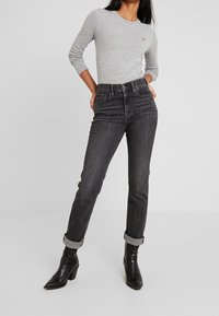 Eine Frau trägt ein graues Langarmoberteil, hoch taillierte, hochgekrempelte schwarze Jeans und schwarze, kniehohe Stiefel mit Absatz und steht mit den Händen in den Taschen.