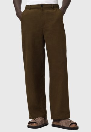 Braune weite Hosen kombiniert mit braunen Sandalen mit offenen Zehen, die Hände in den Taschen, vor einem schlichten hellen Hintergrund.