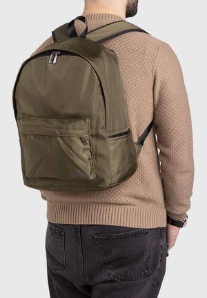 Homme portant un sac à dos vert olive avec poche avant, vêtu d'un pull beige en maille et d'un jean foncé, vu de dos.