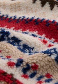 Strukturert fleece-stoff med et gjentakende mønster av røde, blå og beige former på en kremfarget bakgrunn, som fremhever varme og mykhet.
