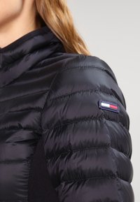 Svart puffjacka med quiltade sektioner och hög krage. Inkluderar en distinkt logotyp i patchform med röd, vit och blå detaljer.