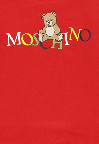 Orsetto marrone seduto sopra il testo colorato "MOSCHINO" su uno sfondo di tessuto rosso.