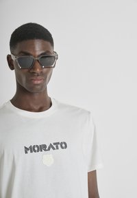 Gafas de sol grises con montura angular, llevadas con una camiseta blanca que presenta el texto negro "MORATO" y un logo de rosa blanca texturizada.