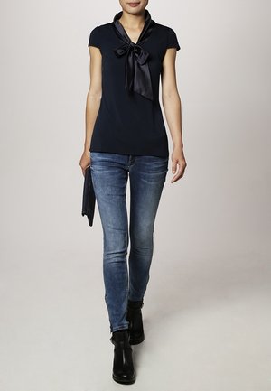 Frau trägt schwarzen kurzärmligen Oberteil mit großer Schleife, blaue Skinny Jeans, schwarze Stiefeletten, hält eine schwarze Clutch, geht nach vorne.