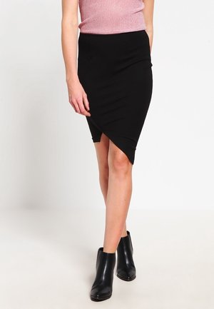 Femme portant une jupe asymétrique noire ajustée, un haut rose sans manches et des bottines noires, debout devant un fond blanc uni.