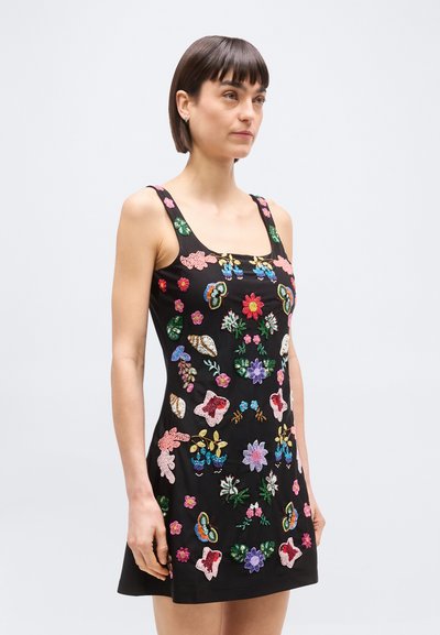 Kvinde iført en ærmeløs sort kjole med farverig blomster- og sommerfuglebroderi, stående mod en ensfarvet lys baggrund.