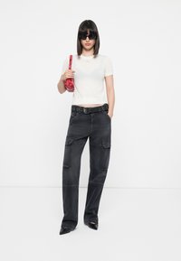 Schwarze Cargo-Hosen mit mehreren Taschen, kombiniert mit einem figurbetonten weißen Kurzarmoberteil. Die Accessoires bestehen aus einer rot gemusterten Tasche und Sonnenbrillen.