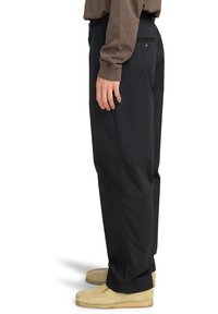 Osoba v tmavých širokých nohaviciach, béžových semišových topánkach a hnedých dlhých rukávoch, stojí v profile na bielom pozadí.