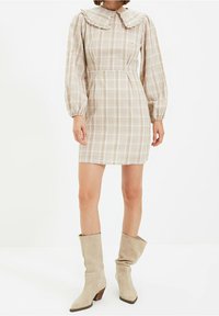 Femme portant une robe mini en plaid beige avec des manches longues bouffantes et un grand col, associée à des bottes en daim beige à tige large.
