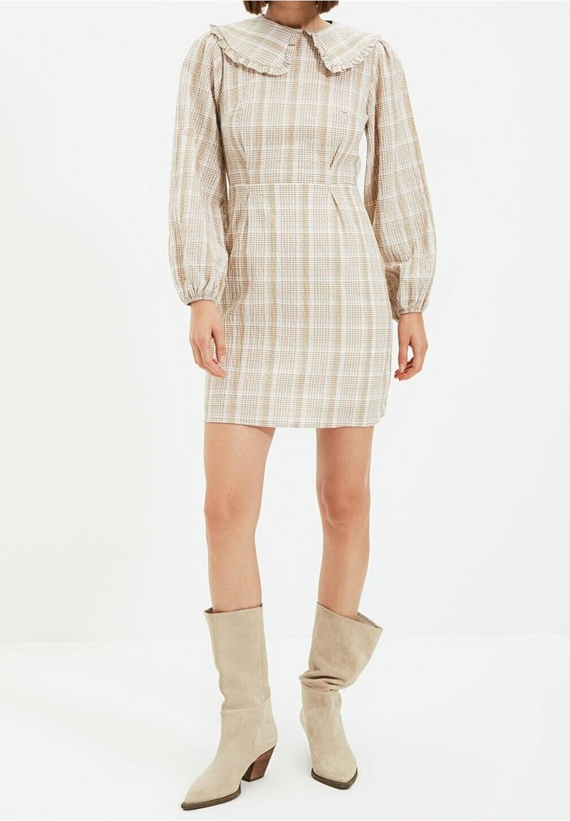 Femme portant une robe mini en plaid beige avec des manches longues bouffantes et un grand col, associée à des bottes en daim beige à tige large.