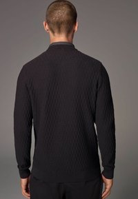 Sort strikket sweater med tekstur, der har krave-nakke, tætsiddende manchetter og et geometrisk mønster gennem hele stoffet, vist fra ryggen.