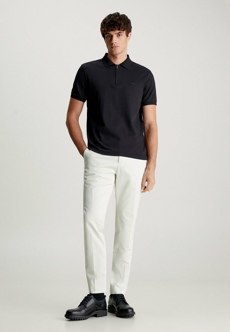 Zwarte polo met een ritskraag, gecombineerd met op maat gemaakte witte broeken. Het model draagt zwarte schoenen en staat op een lichte achtergrond.