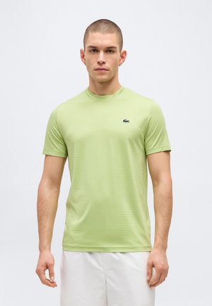 Giovane uomo con capelli corti che indossa una T-shirt Lacoste verde chiaro e pantaloncini bianchi, in piedi davanti a uno sfondo bianco uniforme.
