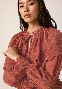 Blouse couleur rouille avec un tissu texturé, broderie florale, encolure à volants et manches bouffantes. Présente un détail de nouage à l'encolure.