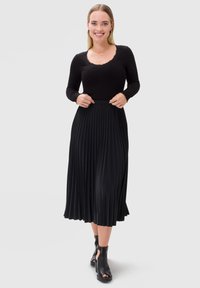 Czarny długi top z długim rękawem i koronkowym dekoltem w połączeniu z plisowaną czarną spódnicą midi. Modelka nosi czarne botki za kostkę z wycięciami.
