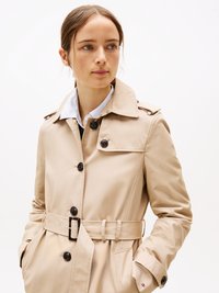 Donna con capelli castano chiaro che indossa un trench beige con cintura, camicia a righe blu e bianca, guarda leggermente di lato.