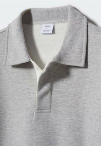 Polo à manches longues gris clair avec col intérieur blanc et une étiquette "MNG" indiquant la taille et le pays de fabrication.