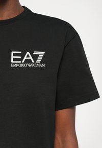 Svart T-skjorte med korte ermer og hvit "EA7 Emporio Armani"-logo på brystet, båret av en person med middels brun hudfarge.