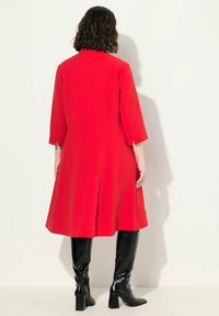 Vestido rojo de longitud media con mangas tres cuartos, una silueta recta y un diseño en A, combinado con botas negras hasta la rodilla.