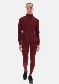 Burgundská bunda na zip s volánovým designem spárovaná s odpovídajícími legínami. Černé tenisky doplňují sportovní outfit. Hladká textura, přiléhavý střih.